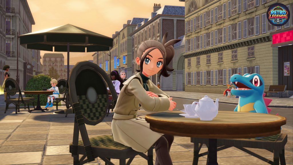 Café-Szene in Pokémon Legenden Z-A: Die Trainerin sitzt mit einem Totodile an einem Tisch im Freien, während im Hintergrund die Sonne über den Dächern von Lumiose City untergeht. Die warme Beleuchtung und die feinen Details an Gebäuden und Geschirr unterstreichen die entspannte Atmosphäre.