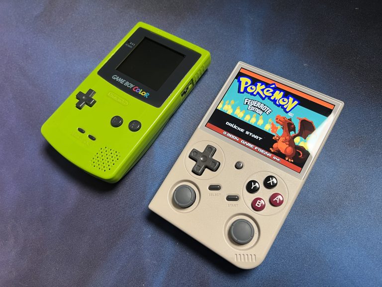 Ein grüner Game Boy Color liegt neben dem einschalteten BATLEXP G350 mit Pokémon Feuerrote Edition auf dem Display. Beide Geräte auf dunkler Oberfläche.