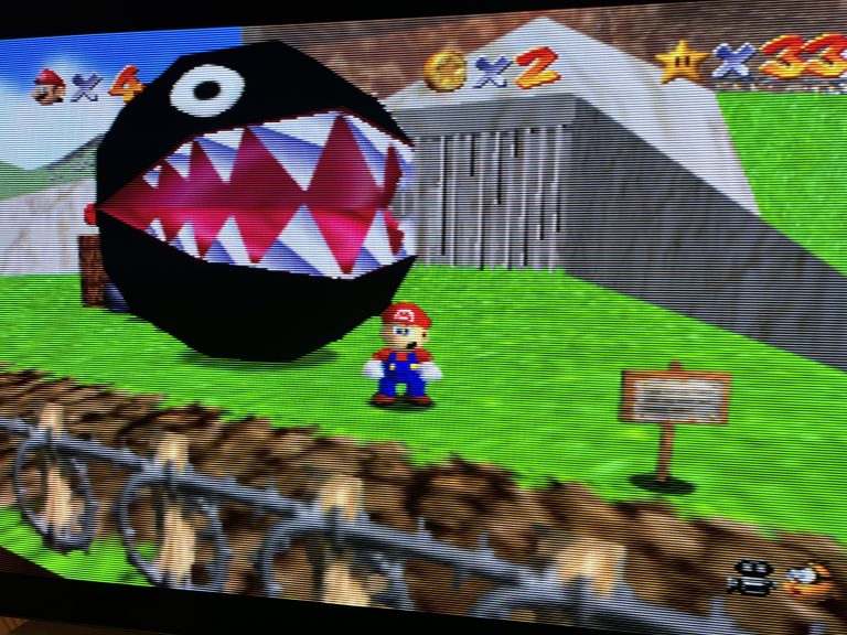 Foto eines TV Bildschirms mit sichtbaren Scanlines. In Super Mario 64 steht Mario auf einer grünen Wiese neben einem riesigen Chain Chomp mit geöffnetem Maul, HUD mit Leben, Münzen und Sternen ist oben eingeblendet.