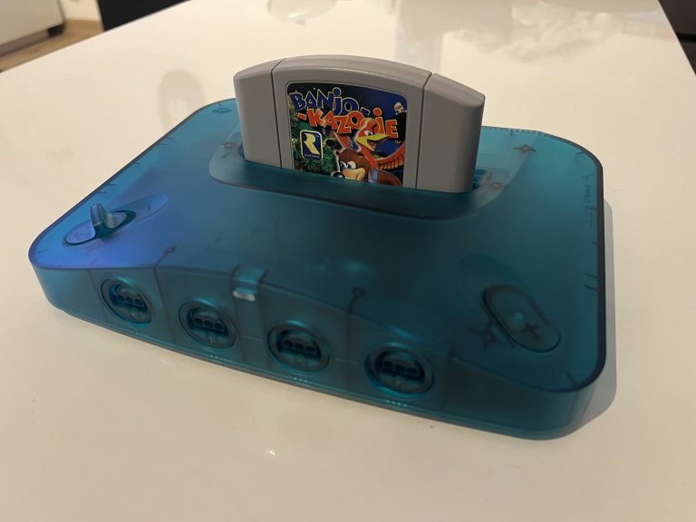 Transparente blaue Analogue 3D Konsole auf einem Tisch, vorne sind vier Controller Anschlüsse sichtbar. Oben steckt ein Banjo Kazooie Nintendo 64 Modul im Cartridge Slot.