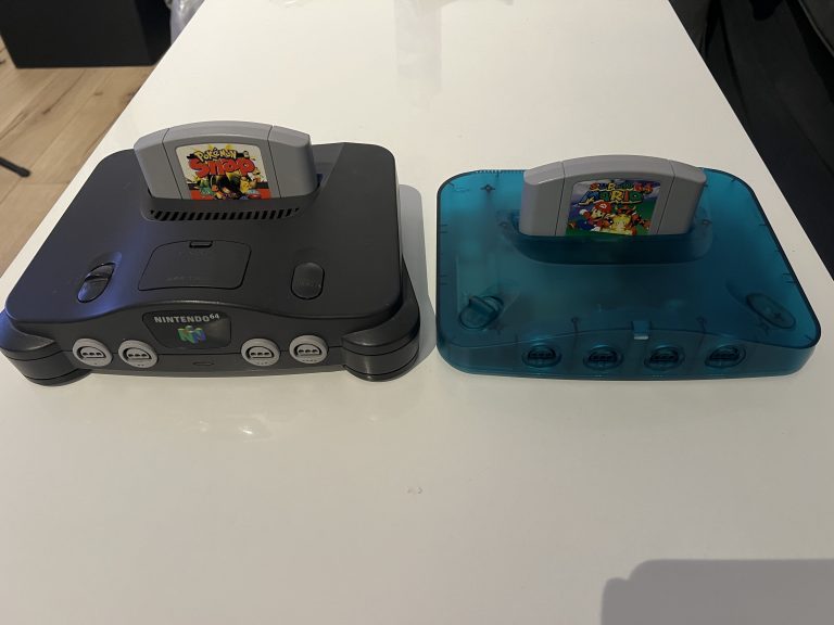 Zwei Konsolen auf einem weißen Tisch, links ein originales Nintendo 64 in dunklem Gehäuse, rechts eine transparente blaue Analogue 3D. In beiden steckt jeweils ein Nintendo 64 Modul, vorne sind die Controller Ports sichtbar.