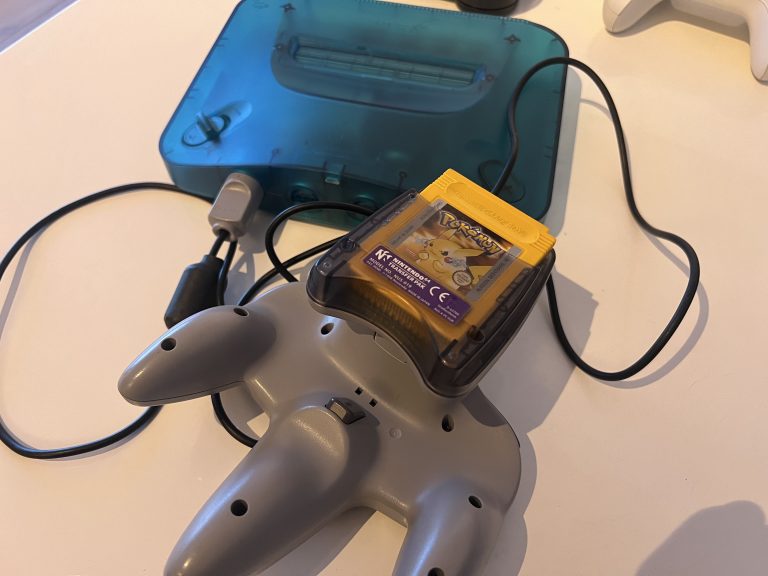Nahaufnahme eines grauen N64 Controllers neben einer transparent blauen Konsole, am Controller steckt ein Transfer Pak mit eingestecktem Pokémon Gelb Game Boy Modul. Kabel liegen locker auf dem Tisch, Setup wirkt wie ein echter Hardware Test.