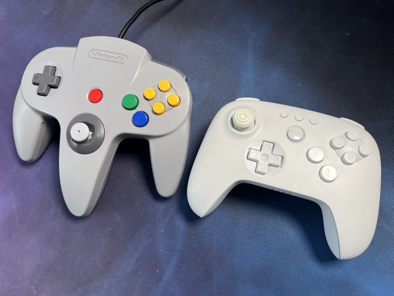 Zwei N64 Controller auf dunklem Untergrund, oben ein originaler grauer Controller mit farbigen Buttons, unten ein helles modernes N64 Pad. Beide sind von oben fotografiert, die Sticks sind gut sichtbar.